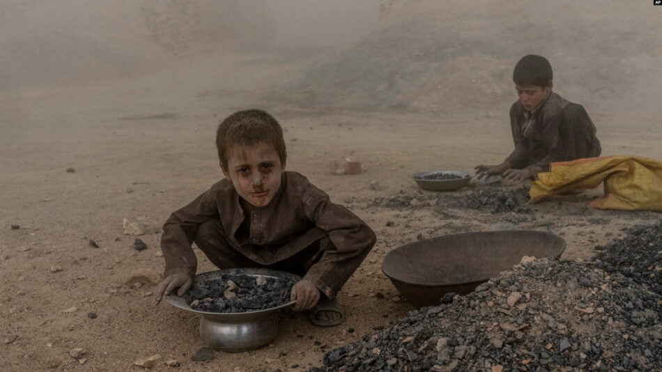 The photo shows two young children sitting in the dirt, collecting half-burnt scraps. Their faces and clothes are dirty, conveying deep despair and poverty.