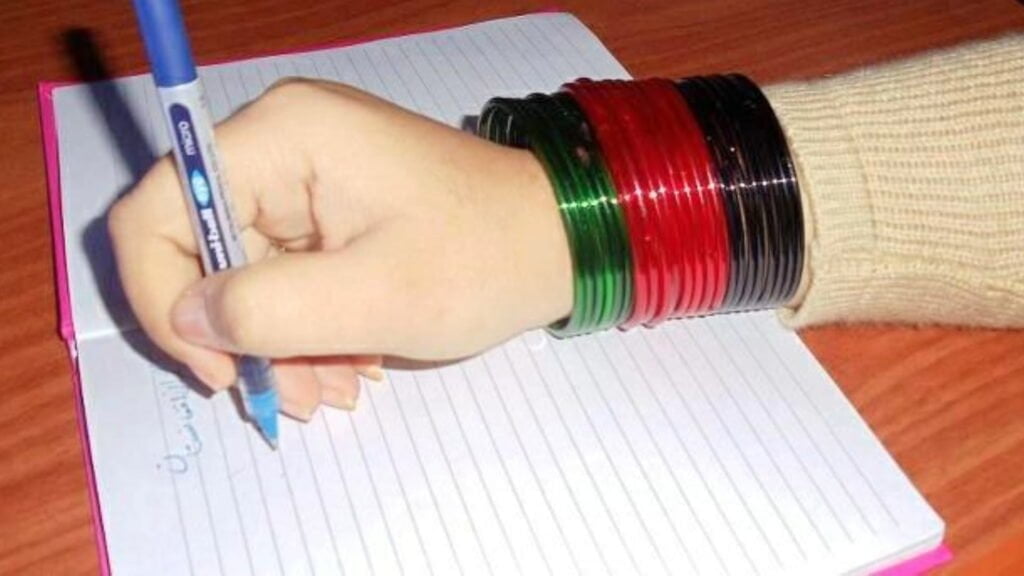 The image shows the hand of a girl writing on paper, wearing bangles that match the colors of the Afghanistan flag