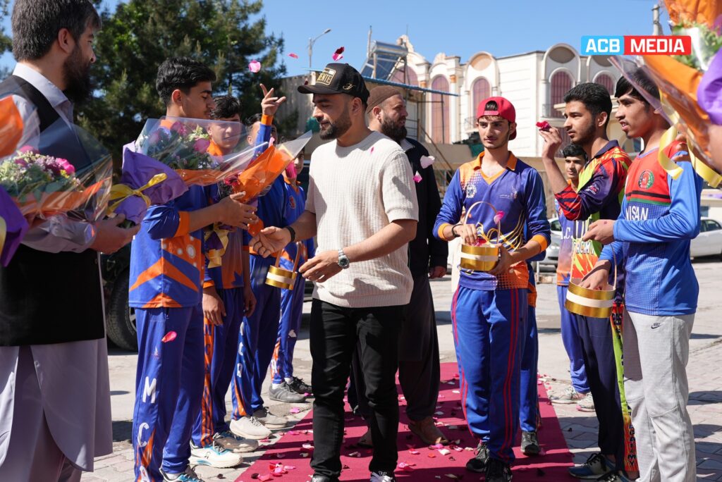 Rashid Khan, the Afghan cricket star, is being warmly welcomed by a group of young cricketers and supporters. He is receiving flower bouquets and standing on a red carpet while being showered with flower petals. The young cricketers, dressed in their colorful uniforms, are gathered around him with smiles and admiration.