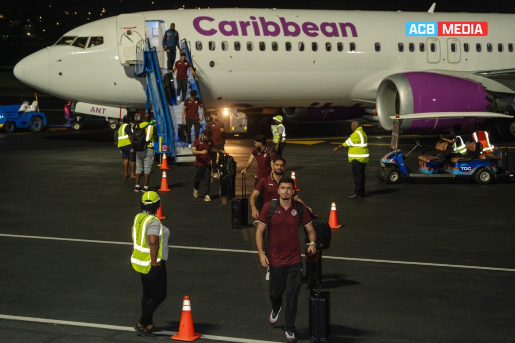 Afghan cricket players have arrived in Saint Kitts and Nevis to participate in the ICC Men's T20 World Cup 2024. The team is seen disembarking from a Caribbean Airlines plane at night, with players carrying their luggage and airport staff guiding them. The players are dressed in maroon team uniforms, ready to compete in the tournament