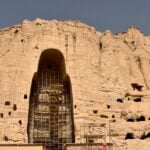 Scaffolding surrounds the remains of a large Buddha statue carved into the cliffside in Bamiyan, Afghanistan. The empty niche is all that remains after the statue was destroyed, with the surrounding rocky landscape dotted with smaller caves and holes under a clear blue sky.