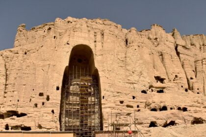 Scaffolding surrounds the remains of a large Buddha statue carved into the cliffside in Bamiyan, Afghanistan. The empty niche is all that remains after the statue was destroyed, with the surrounding rocky landscape dotted with smaller caves and holes under a clear blue sky.
