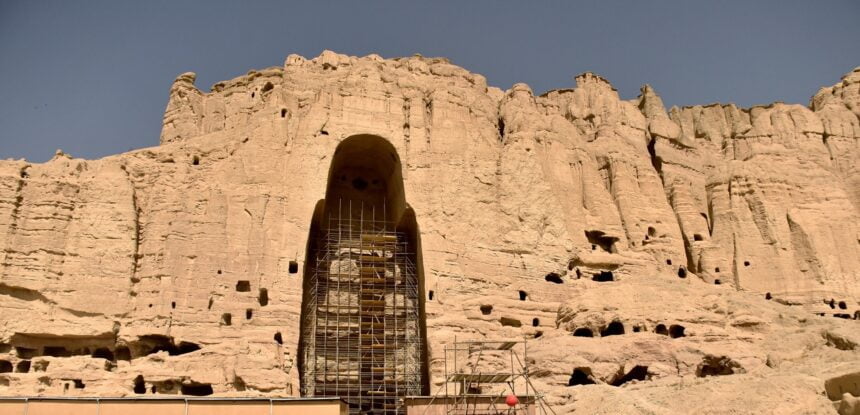 Scaffolding surrounds the remains of a large Buddha statue carved into the cliffside in Bamiyan, Afghanistan. The empty niche is all that remains after the statue was destroyed, with the surrounding rocky landscape dotted with smaller caves and holes under a clear blue sky.