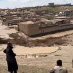 Floodwaters inundate a village in Ghor, Afghanistan, causing severe damage to homes and infrastructure. Residents observe the destruction from a distance.