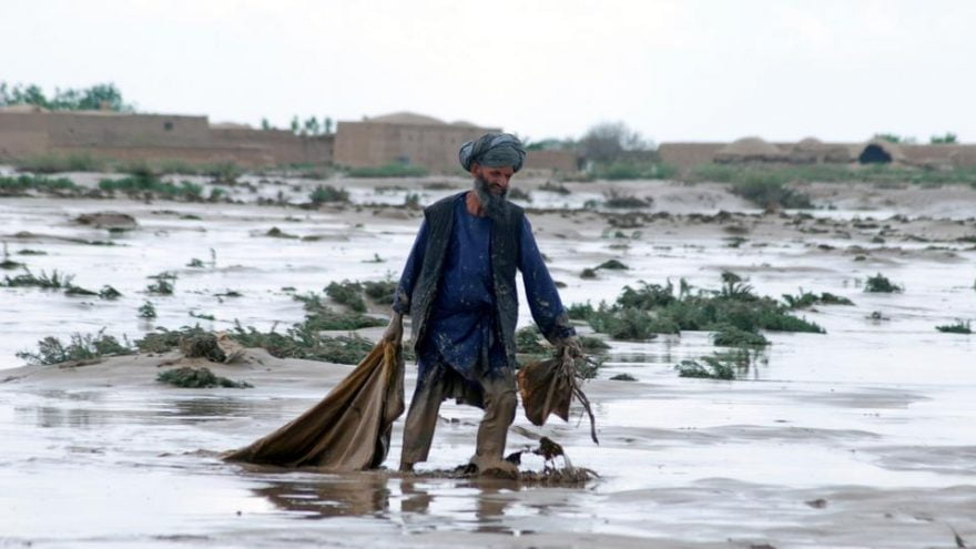 An Afghan man walks through a flooded field, carrying a sack and navigating through water and debris, with sparse vegetation and mud-covered ground visible around him and traditional mud houses in the background.