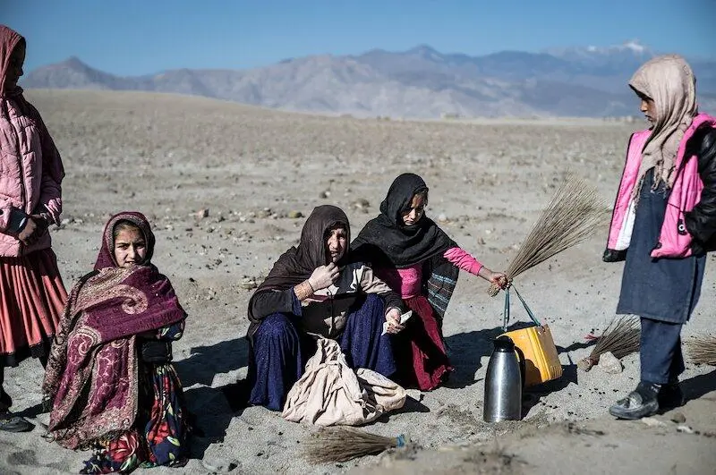 Afghan women and children gathered in a drought-stricken, arid landscape. One elderly woman sits wrapped in a shawl, while two younger women, one handing a broom to a child, are seated on the ground. A young girl in a pink jacket stands to the side, observing. The environment is barren with sparse vegetation and distant mountains under a clear sky, highlighting the harsh living conditions