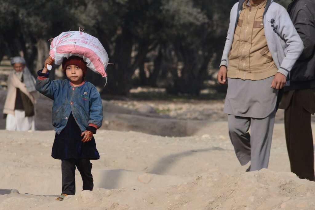 A small Afghan girl, her face set with quiet determination, balances a sizable sack on her head while navigating a dusty, uneven path. An older boy follows behind her, their surroundings hinting at hardship and scarcity.