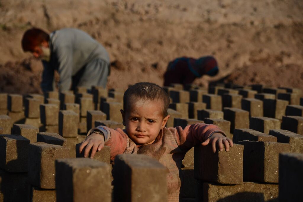 A very young Afghan child stands amid rows of clay bricks at a makeshift kiln, their face and clothes smeared with dust. In the background, an adult figure works on shaping more bricks, highlighting the arduous conditions and early introduction to labor.