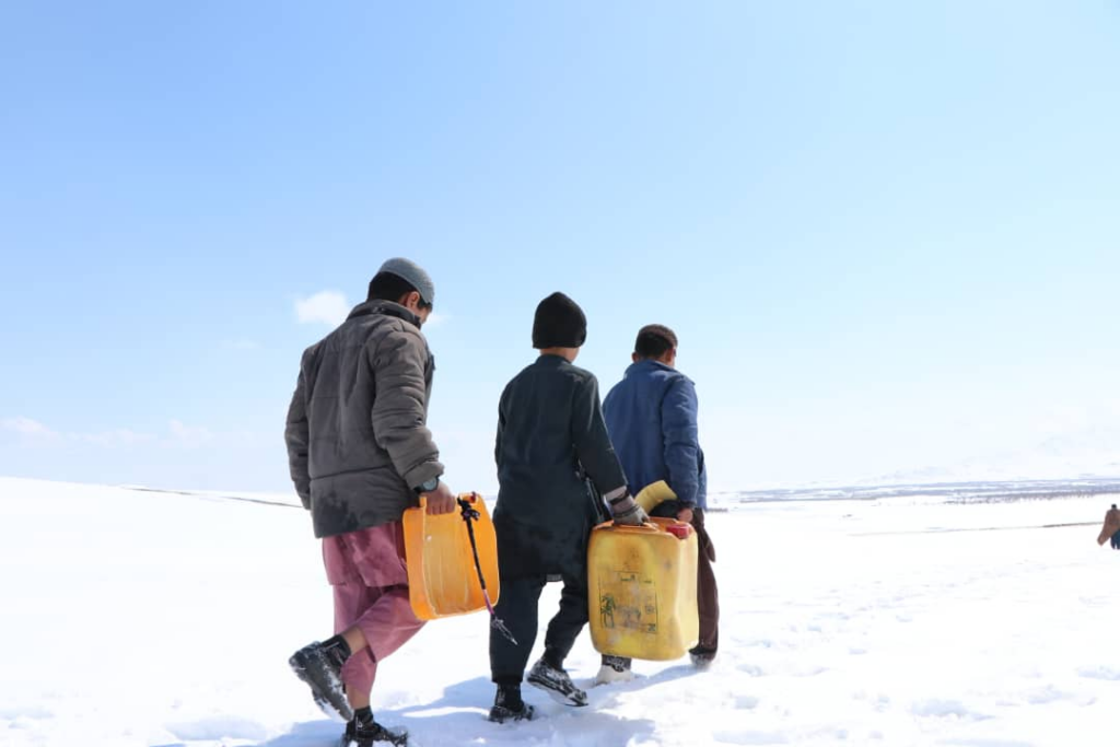 Three young Afghan boys walk across a snow-covered landscape under a bright blue sky. Each carries a large yellow plastic jerrycan, their winter clothing and body language reflecting the effort of moving through the cold, white terrain.